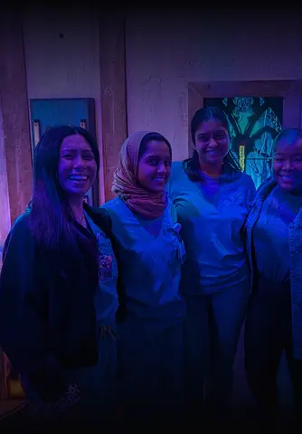 Four players smiling in medical scrubs inside an immersive room at Huddy's Escape, lit with blue and purple ambient lighting.
